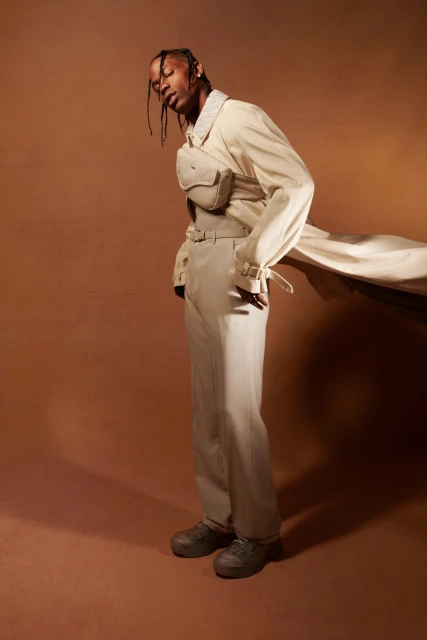 Man in cream trench coat and pants stands angled on mocha backdrop, neutral tones, studio lighting, fashion shoot with modern styling.