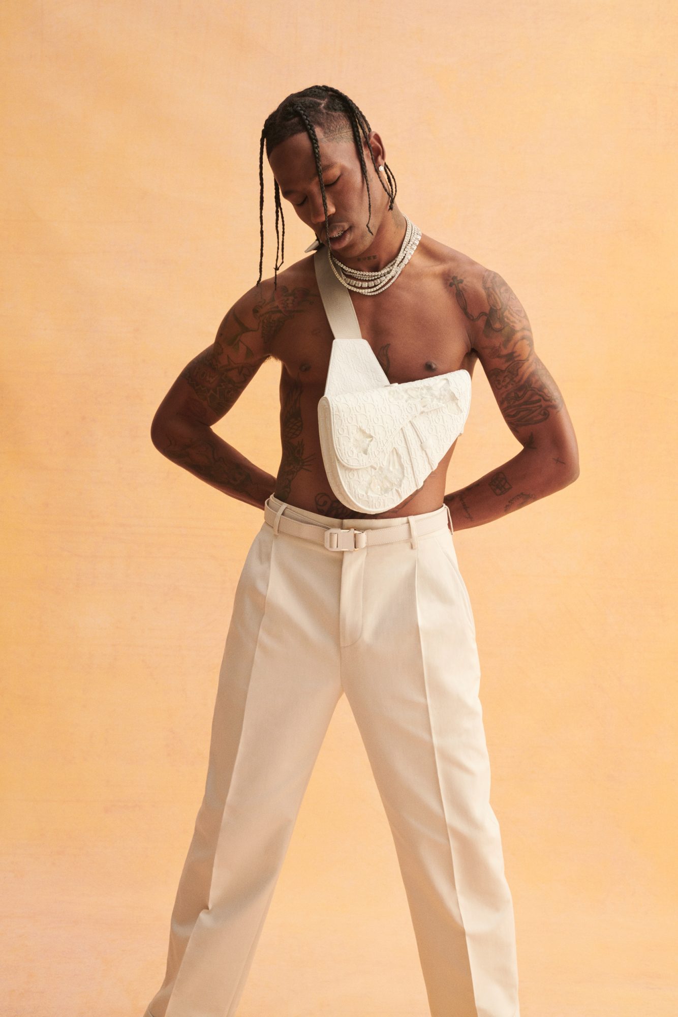 A shirtless adult man poses in a studio wearing white pants, jewelry, and a white textured belt bag against an orange gradient background.