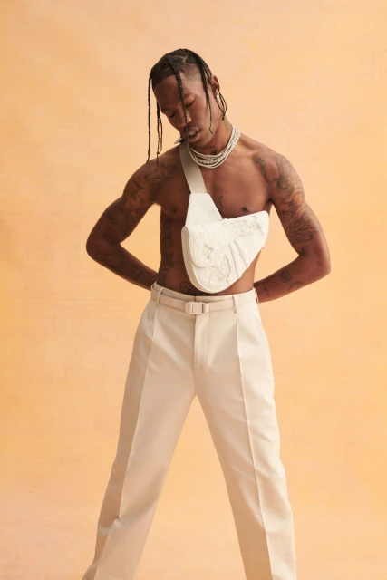 A shirtless adult man poses in a studio wearing white pants, jewelry, and a white textured belt bag against an orange gradient background.