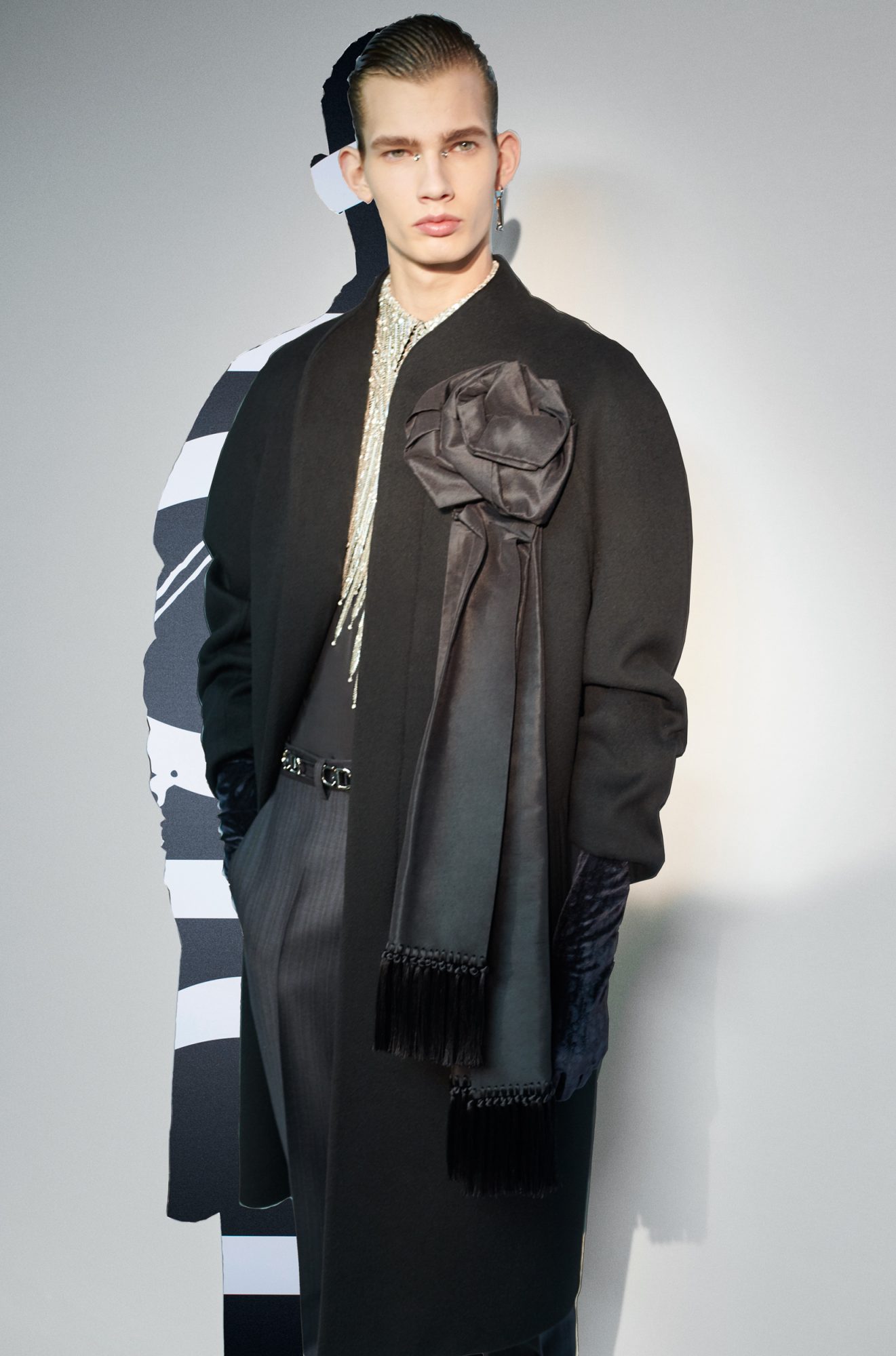 Male fashion model poses in a long black coat with a large fabric flower and shiny fringe detail, standing indoors against a gray background.