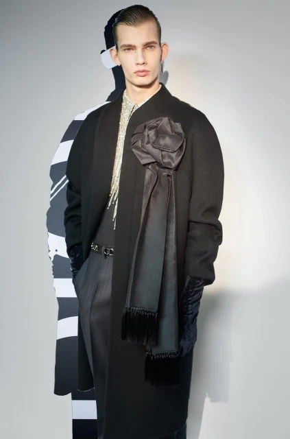 Male fashion model poses in a long black coat with a large fabric flower and shiny fringe detail, standing indoors against a gray background.