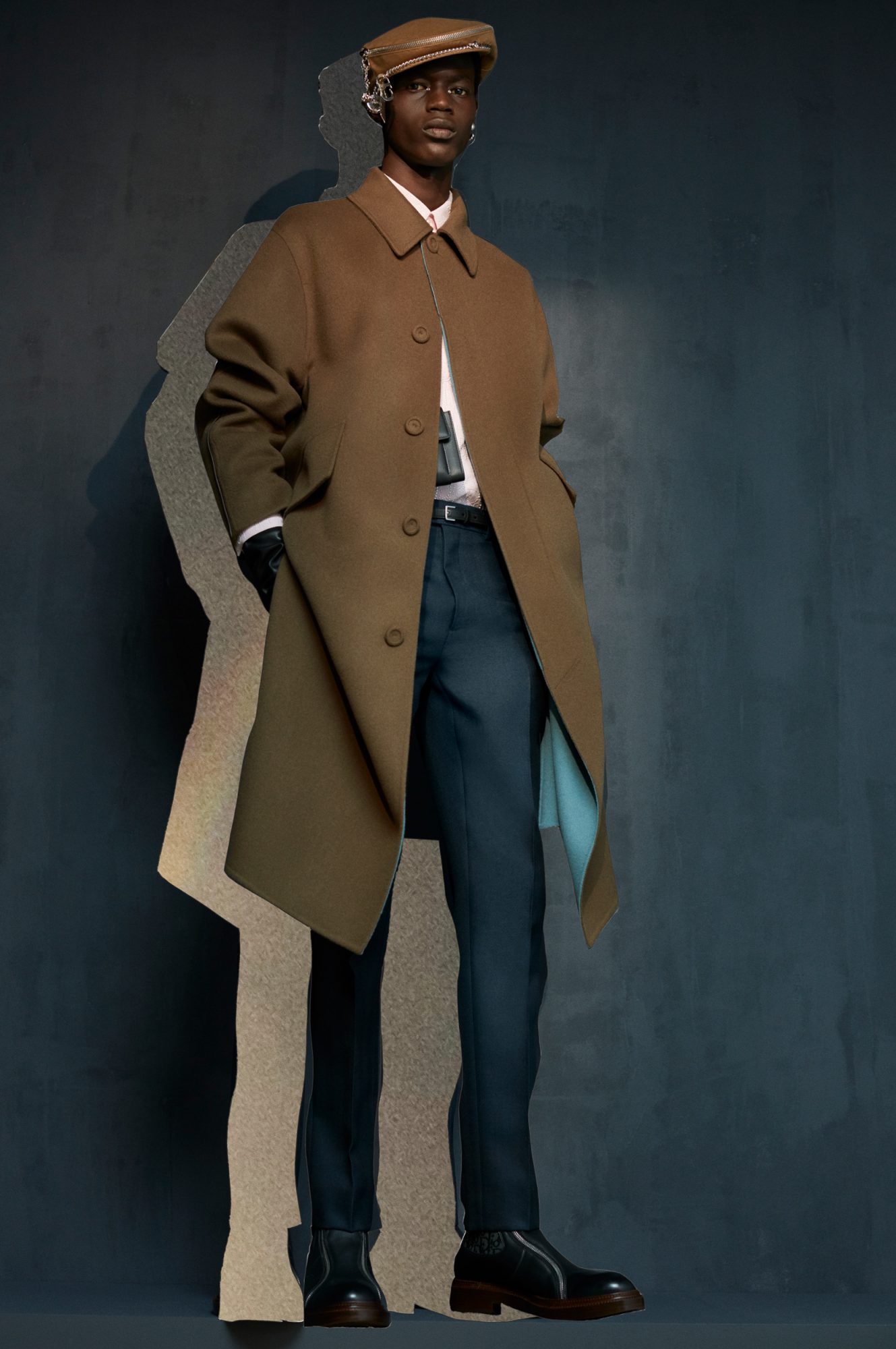 A stylish Black male model poses in a brown overcoat, white collared shirt, and dark blue pants against a blurred grey-blue backdrop.
