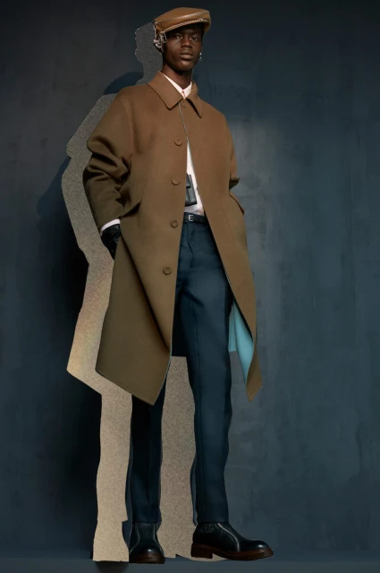 A stylish Black male model poses in a brown overcoat, white collared shirt, and dark blue pants against a blurred grey-blue backdrop.