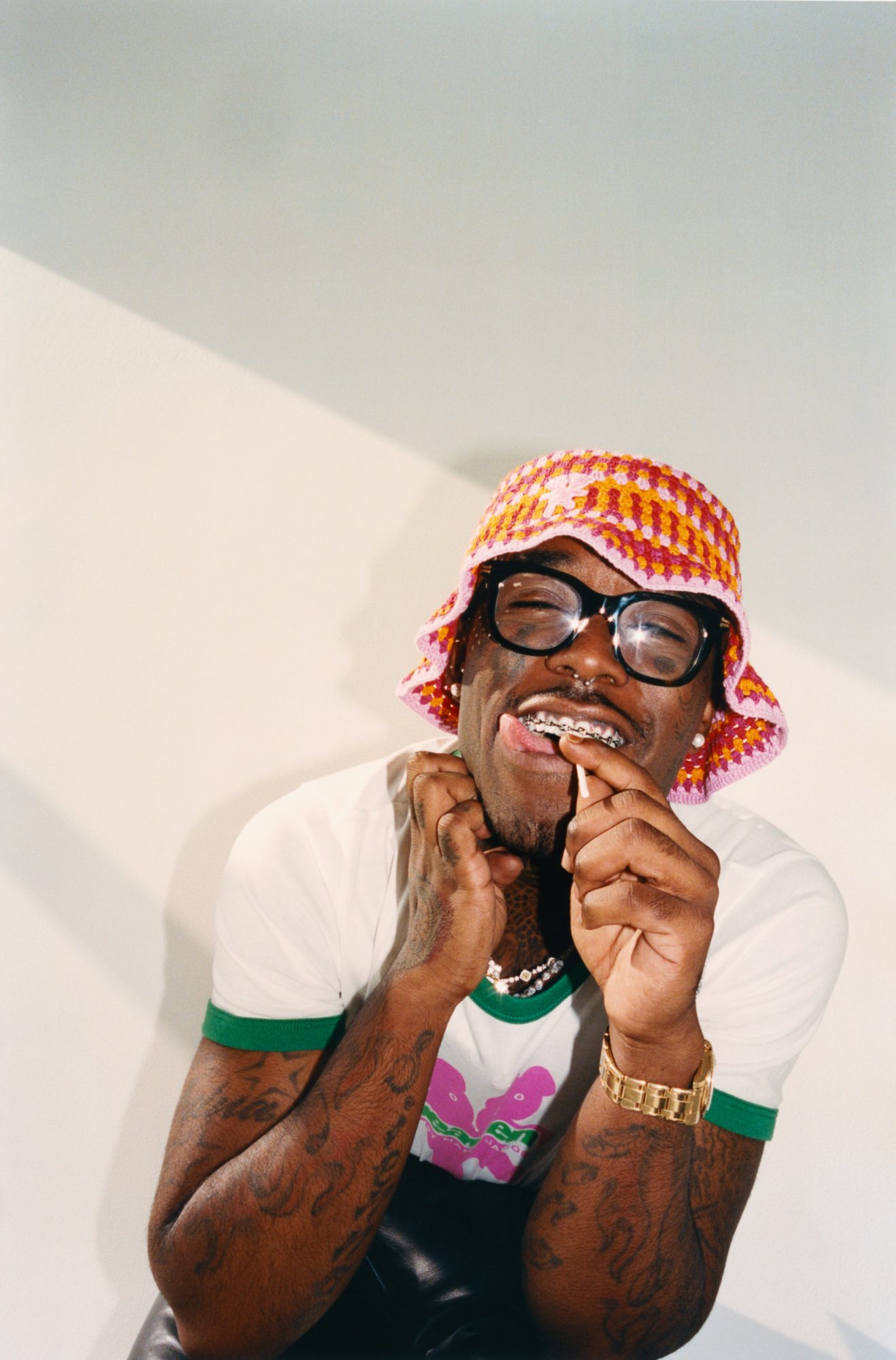 Portrait of a person wearing a colorful crochet hat, black glasses, a white shirt with green trim, and jewelry with braces.
