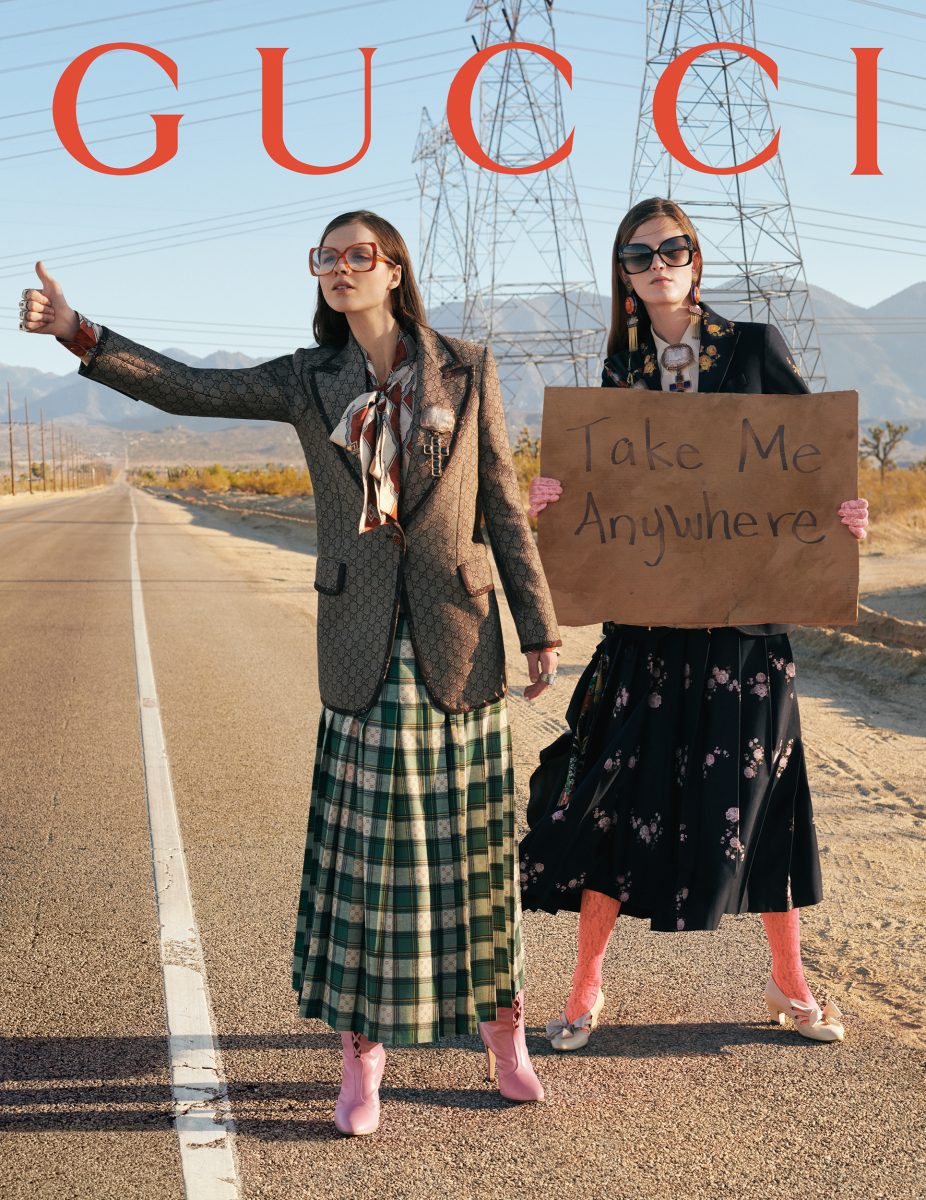 Two women stand on a desert road with power lines in background. One holds a 'Take Me Anywhere' sign in this fashion editorial.