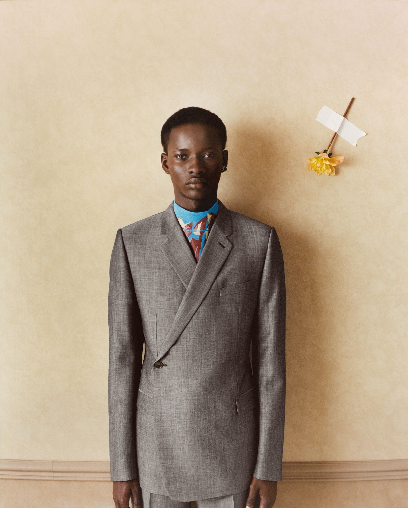 Portrait of a Black male model wearing a grey suit jacket and colourful shirt, standing against a neutral wall with a flower taped to it.