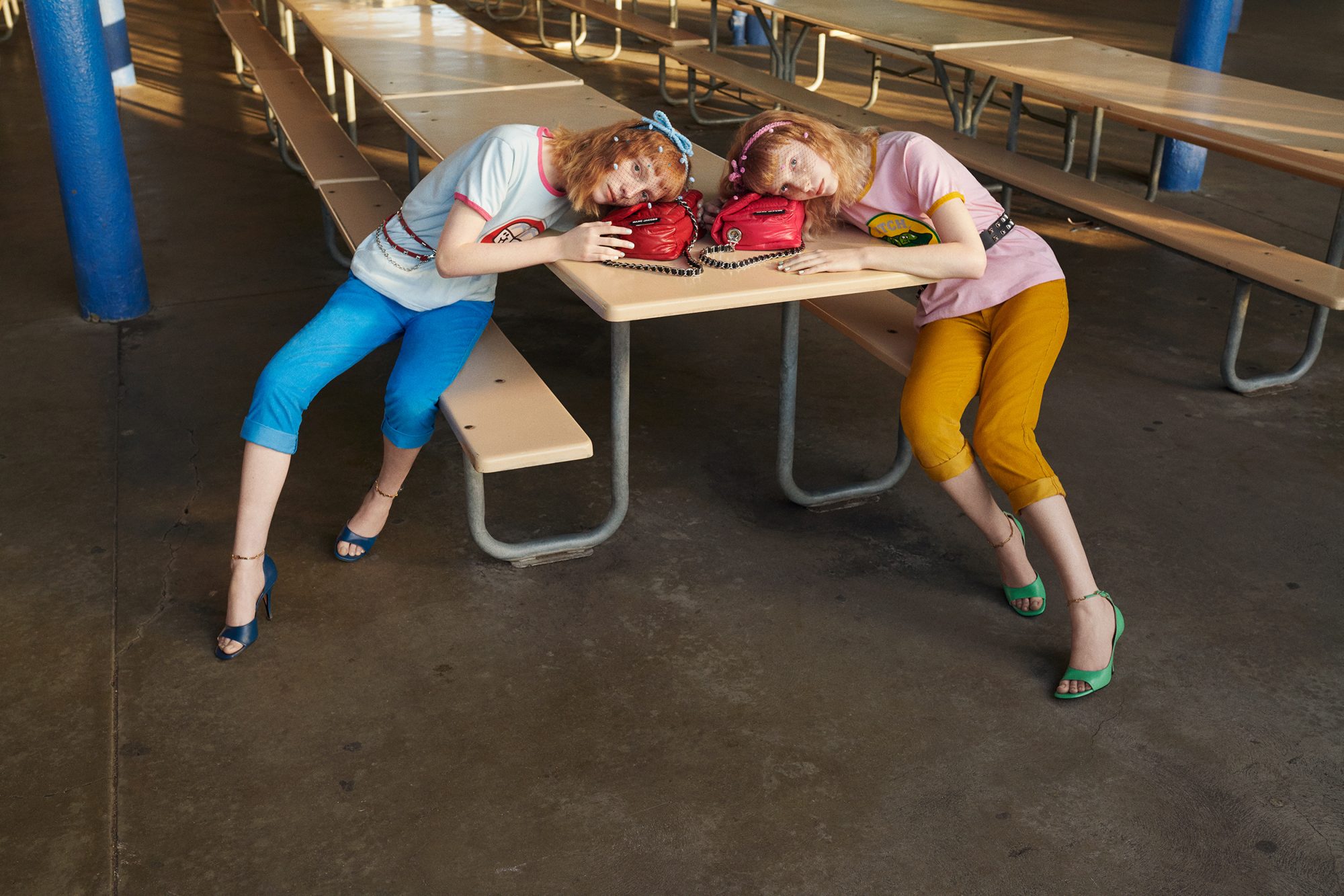Two young women pose at a picnic table, showcasing red handbags, with blue and yellow outfits in an outdoor setting. Fashion editorial shot.