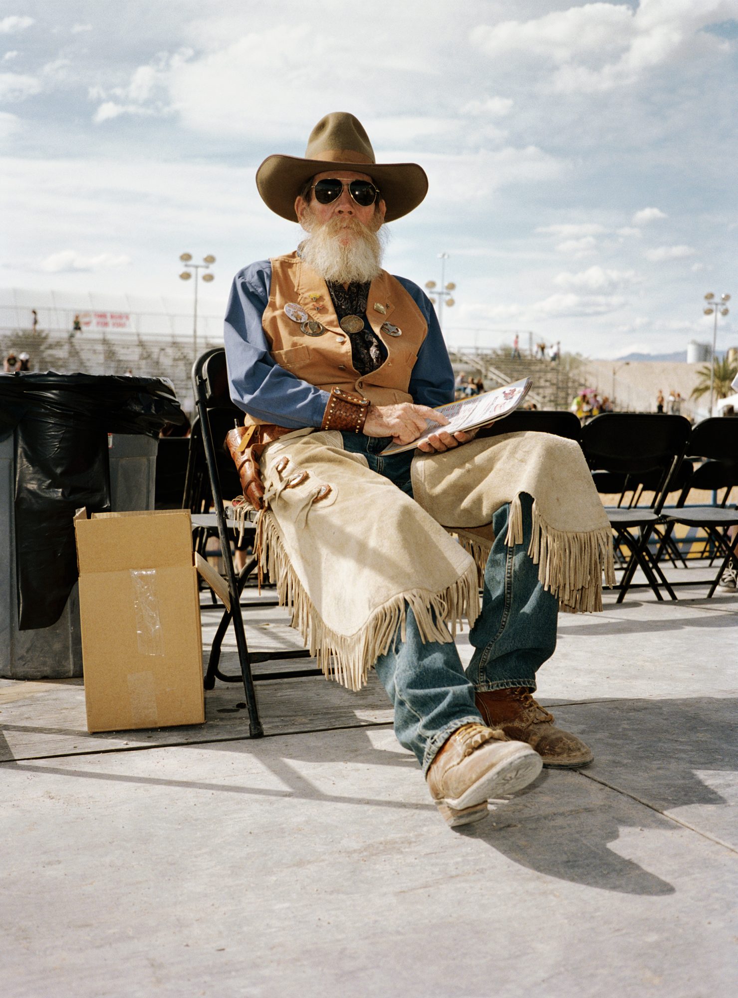 An elderly man dressed in cowboy attire sits in a folding chair outdoors on a sunny day, holding a program, and wearing sunglasses.
