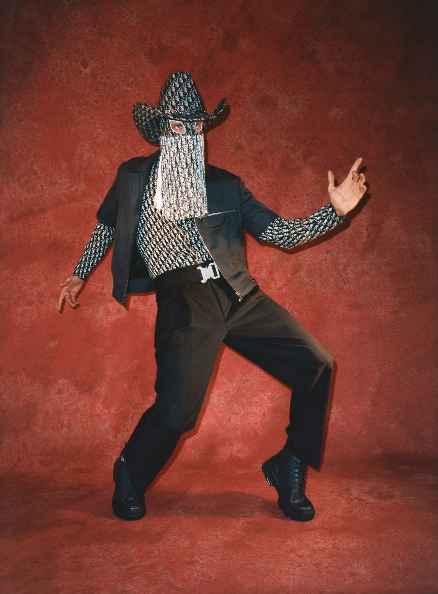 Full-length studio shot of a person in a black suit, cowboy hat, and mask with a designer print, striking a pose against a red backdrop.