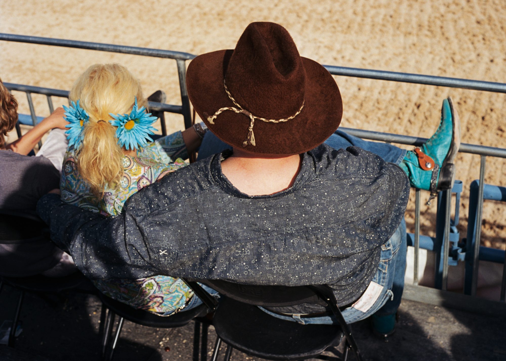 Overhead shot of three people wearing cowboy boots, hats, and jeans sitting on a metal railing next to a sandy outdoor space.