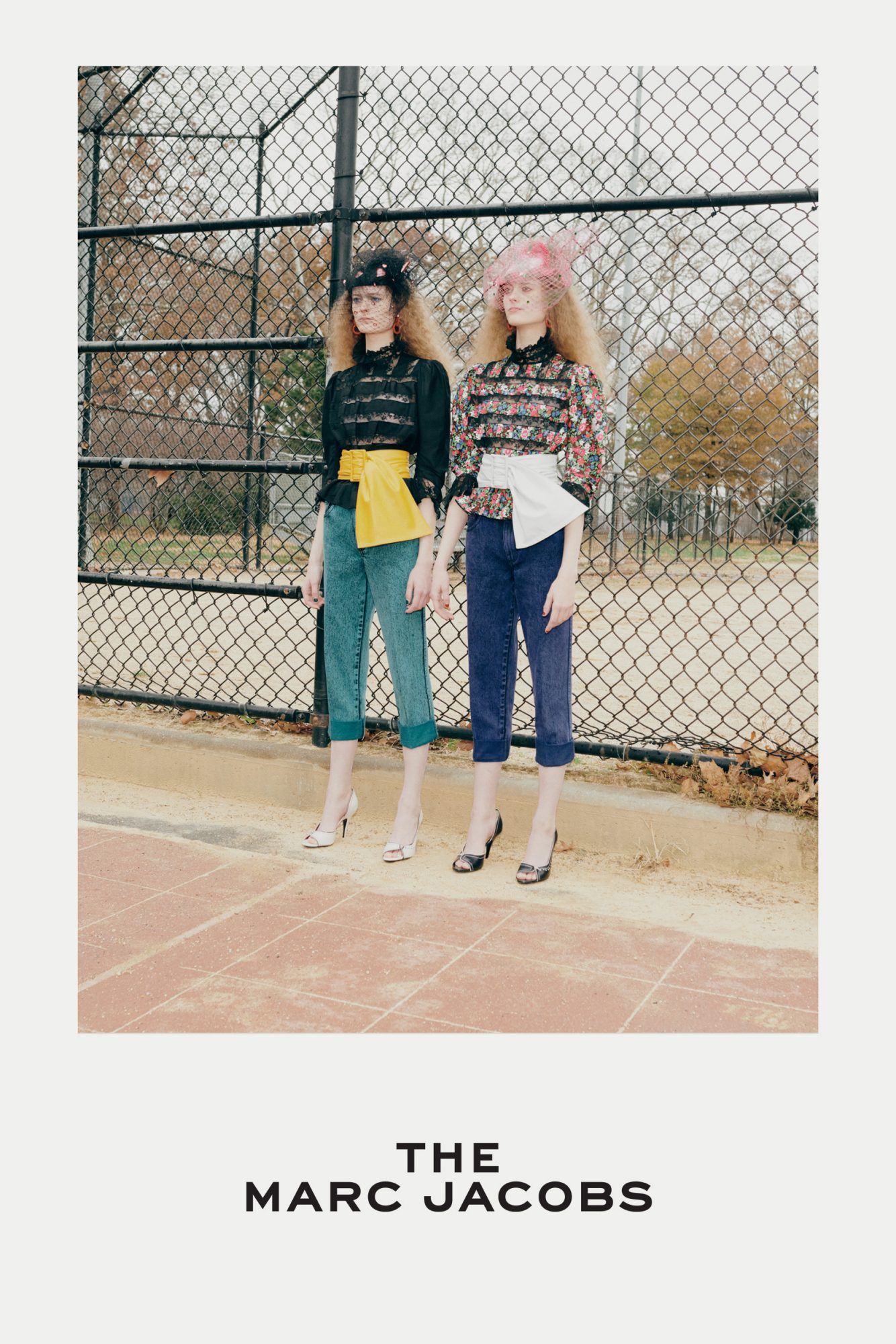 Two fashion models pose on a tennis court in front of a chain link fence, wearing vintage-inspired outfits with hats and high heels.