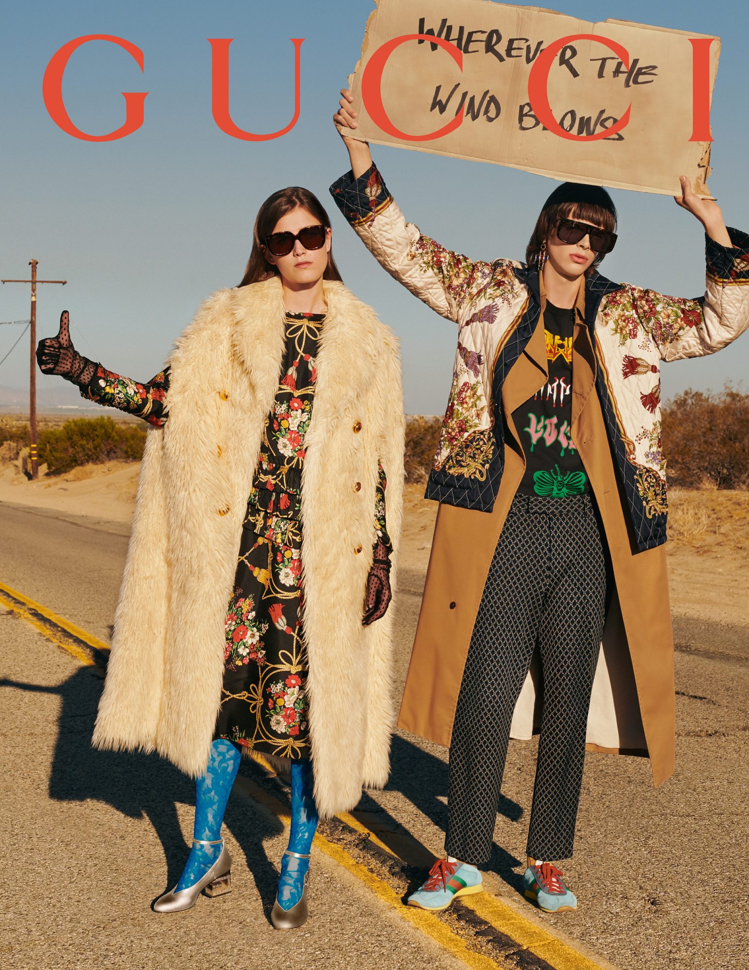 Two fashion models stand on a desert road holding a sign for a Gucci advertisement, wearing oversized outerwear and tinted glasses.