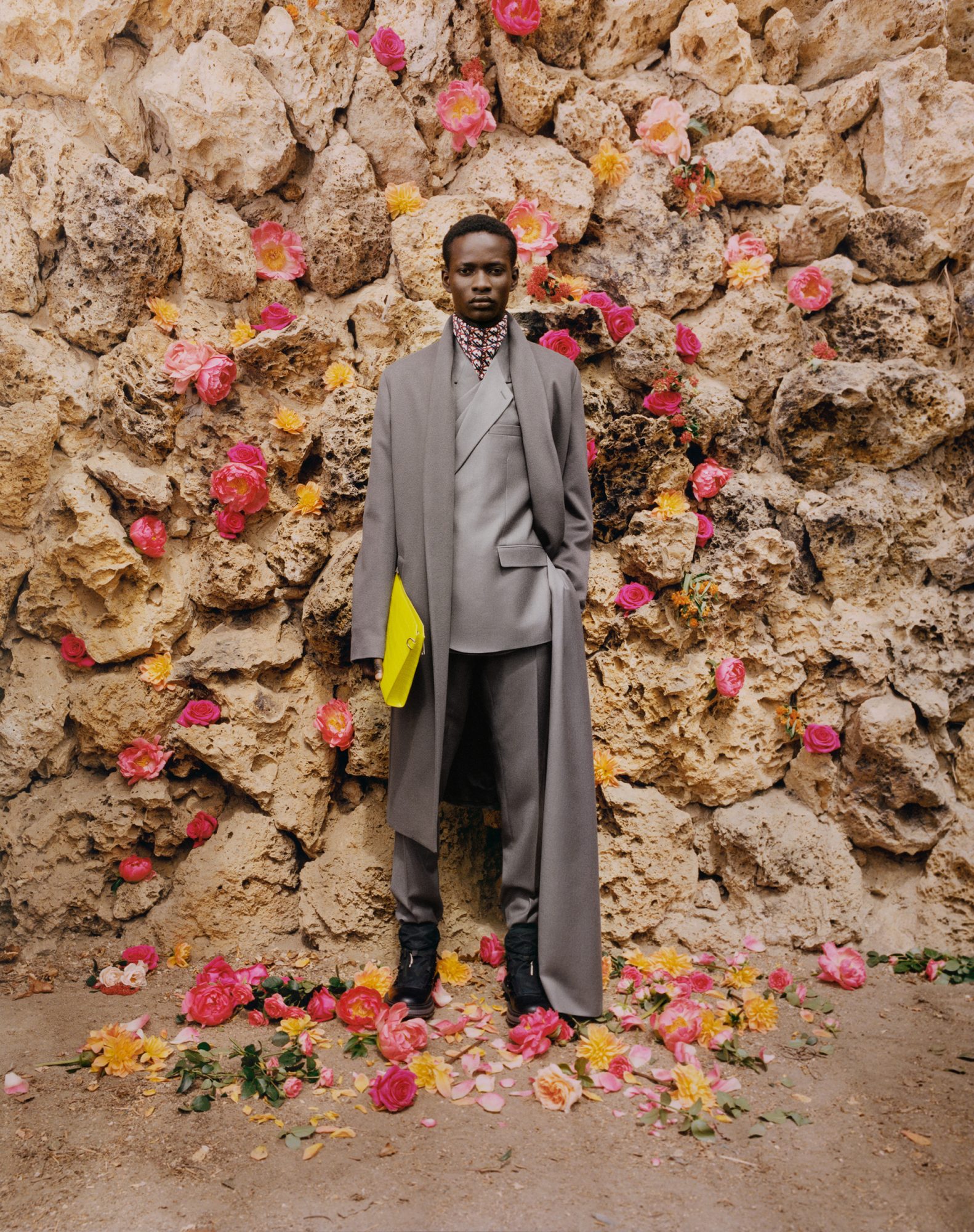 An adult stands outdoors wearing a grey suit and overcoat, holding a yellow folder, posing in front of a stone wall covered with flowers.