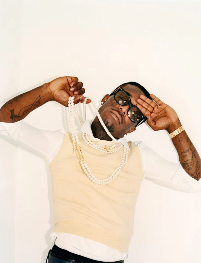 Black man wearing sunglasses and layered pearl necklaces holds multi-strand pearls against a white background