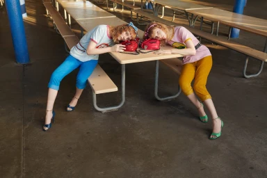 Two red-haired models resting heads on a picnic table wearing colorful outfits photographed by Hugo Scott