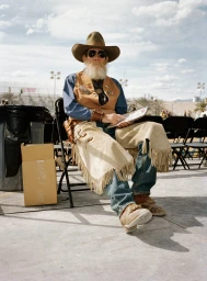 Elderly man in cowboy hat and sunglasses seated outdoors, wearing a tan leather vest and fringed chaps over denim.