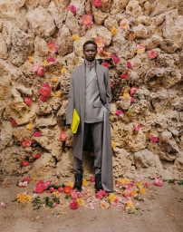 A man in a grey suit and long coat holding a bright yellow pouch against a stone wall embedded with pink and yellow flowers.