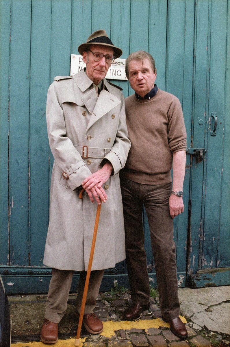 Two men stand side-by-side in front of a blue wooden door. The man on the left is older, wearing a trench coat and hat.