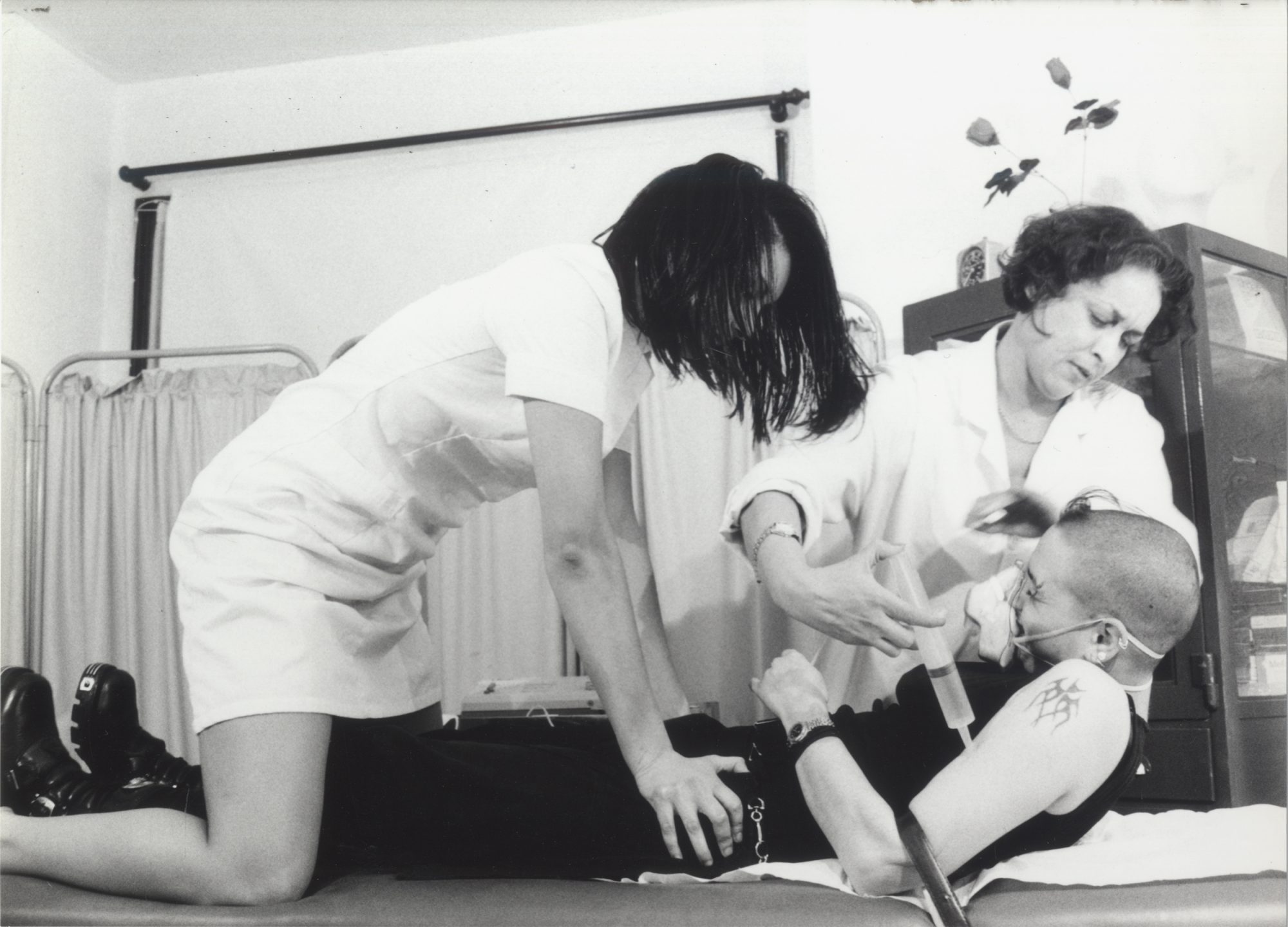 Medical scene showing a patient lying down while two medical professionals attend to them, set in a clinical indoor environment.