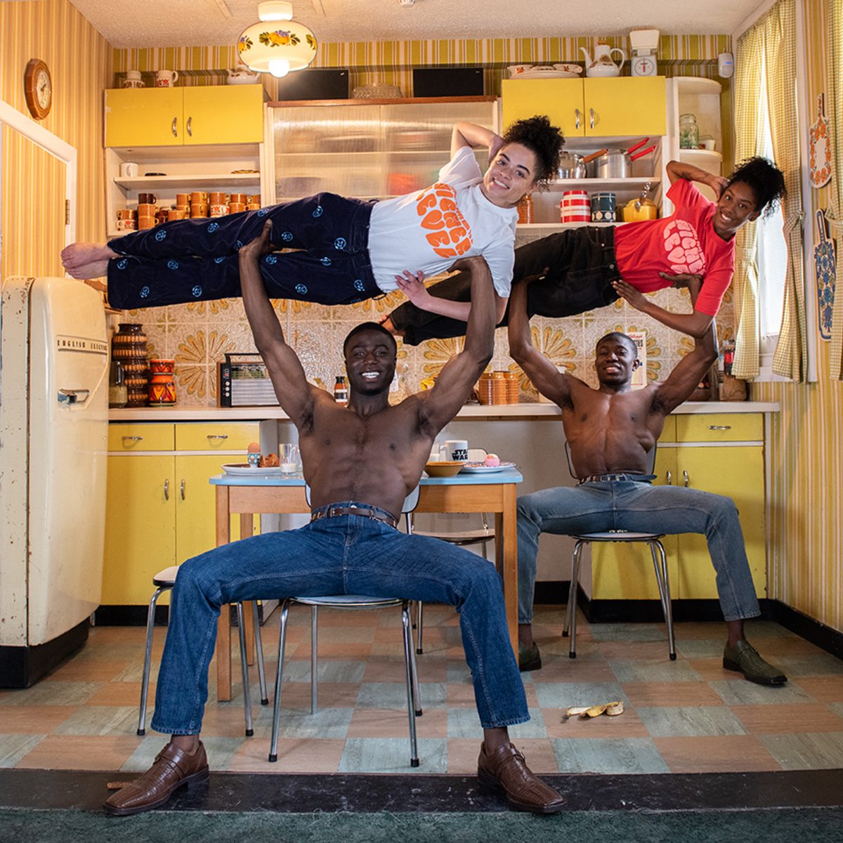 Two men lift women in a playful kitchen scene featuring retro decor, bright colors, and dynamic poses, blending strength and humor indoors.
