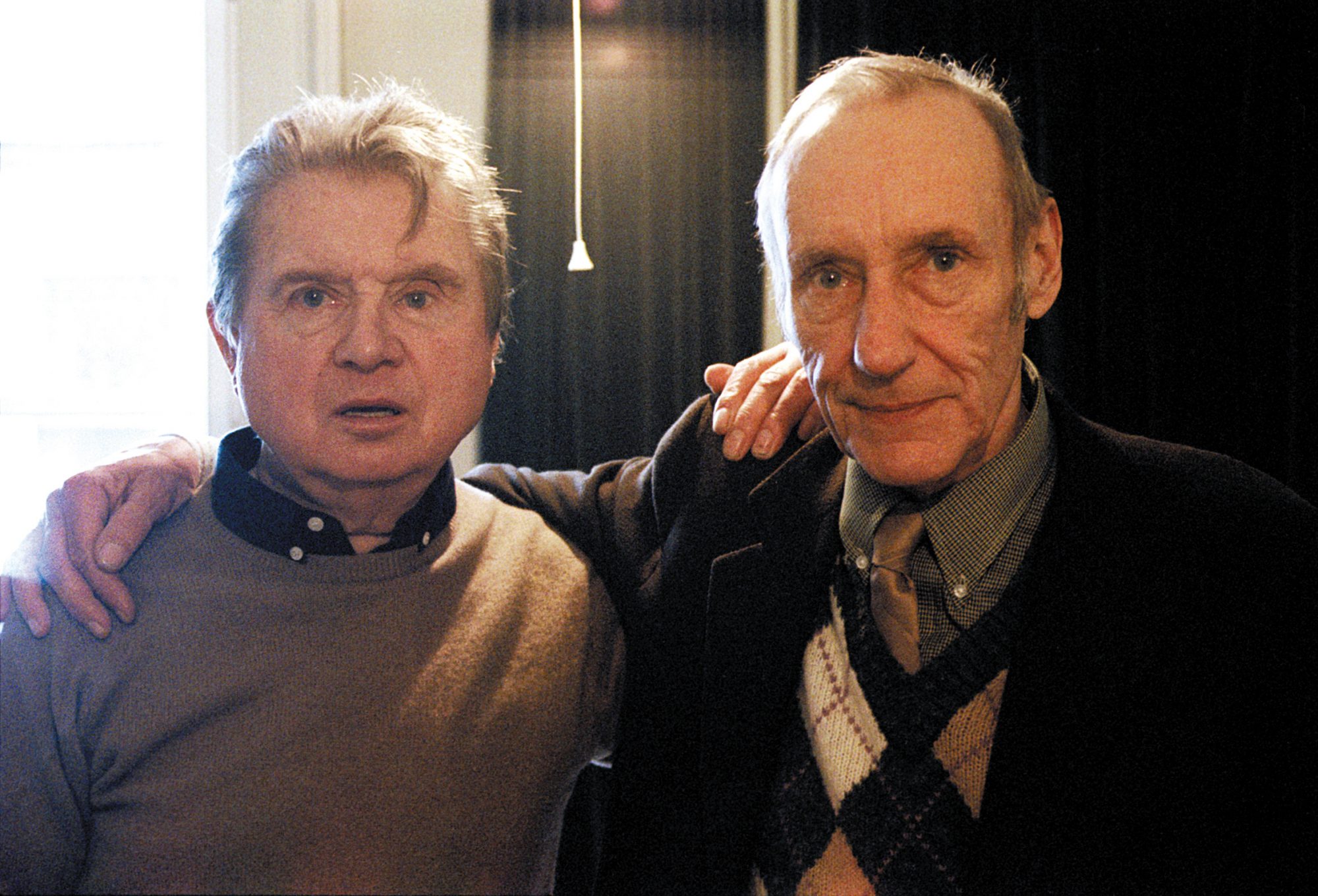 Two adult males stand indoors in front of a window. One man has his arm around the other, wearing a sweater and jacket.