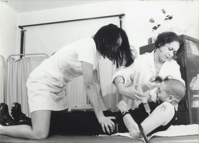 Black and white photograph by Dick Jewell of three women in a medical setting, one person receiving an injection on a table.