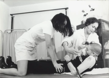 Black and white photograph by Dick Jewell of three women in a medical setting, one person receiving an injection on a table.
