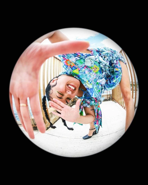 A fisheye shot of a woman in a floral dress smiling with her hands raised on a balcony overlooking the ocean against a clear sky.