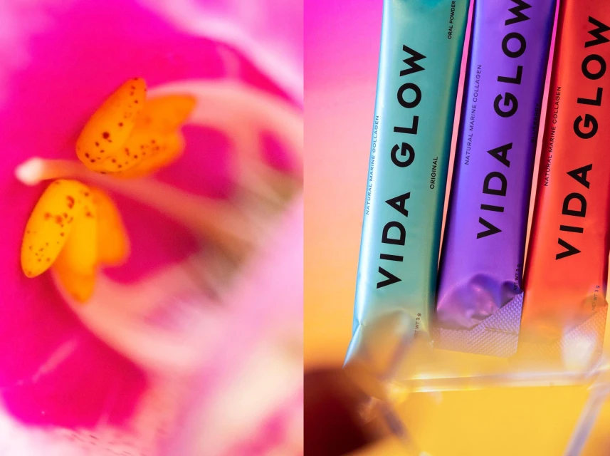 Close-up of three 'Vida Glow' supplement sticks in teal, purple, and red, displayed on a reflective surface, alongside pink flower.