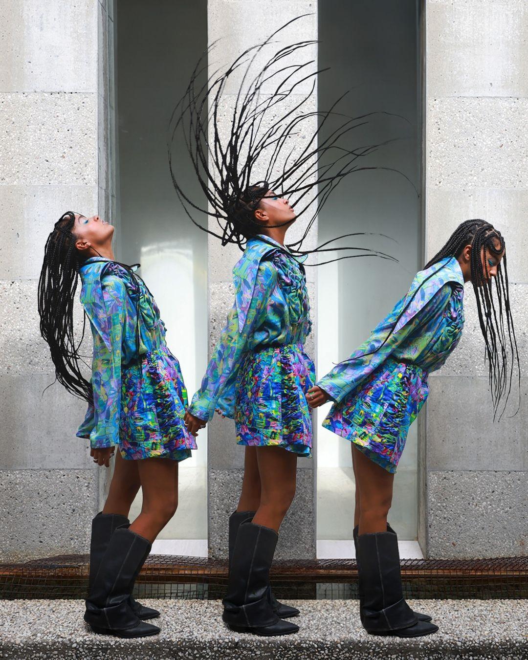 Three adult females in patterned dresses and tall boots stand together holding hands in front of a neutral stone wall and floor.