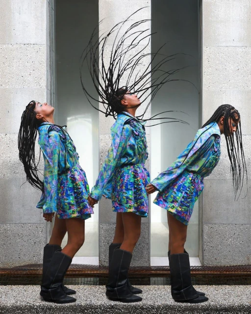 Three adult females in patterned dresses and tall boots stand together holding hands in front of a neutral stone wall and floor.