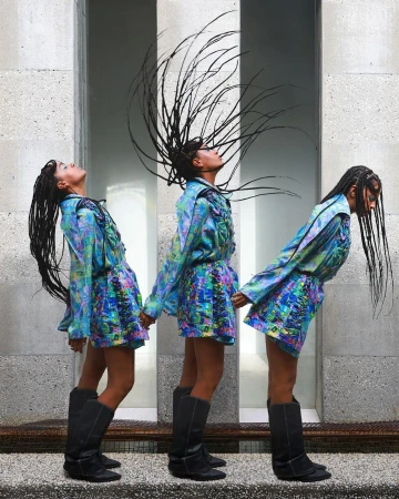 Three adult females in patterned dresses and tall boots stand together holding hands in front of a neutral stone wall and floor.