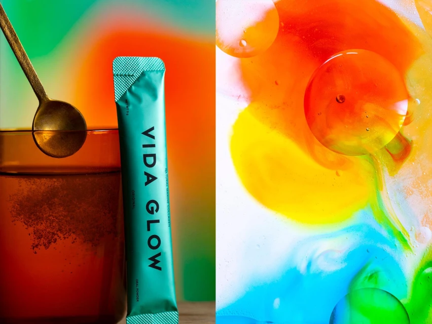 A glass with water, a gold spoon, and a teal Vida Glow supplement packet against an abstract background with bright colors.