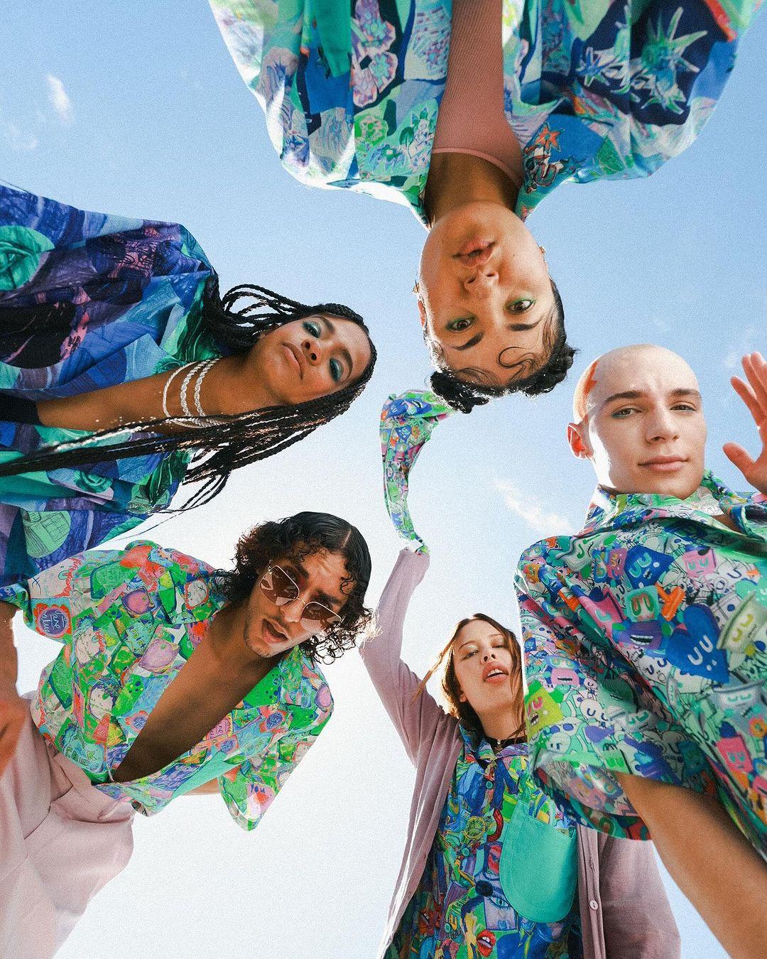 Group of five diverse young adults in patterned shirts, looking down into camera from low angle under bright blue sky on sunny day.