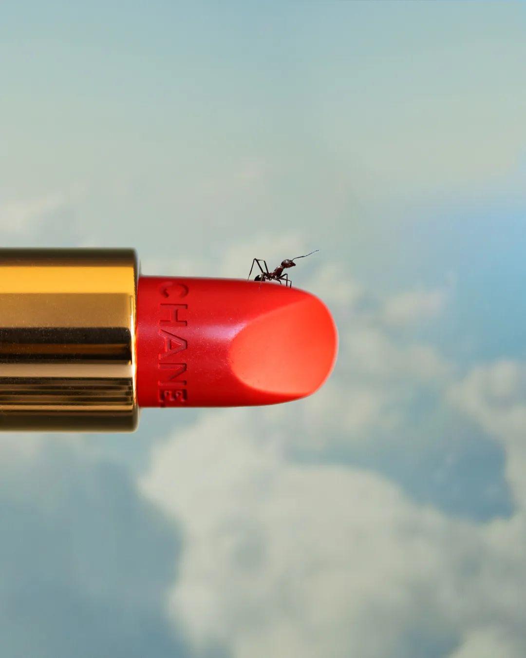 A red Chanel lipstick tube with a gold base and an ant perched on top, set against a blurred blue sky with fluffy white clouds.