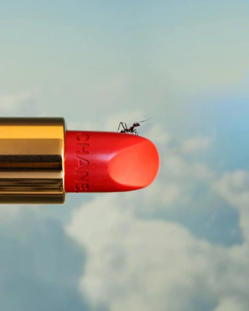 A red Chanel lipstick tube with a gold base and an ant perched on top, set against a blurred blue sky with fluffy white clouds.