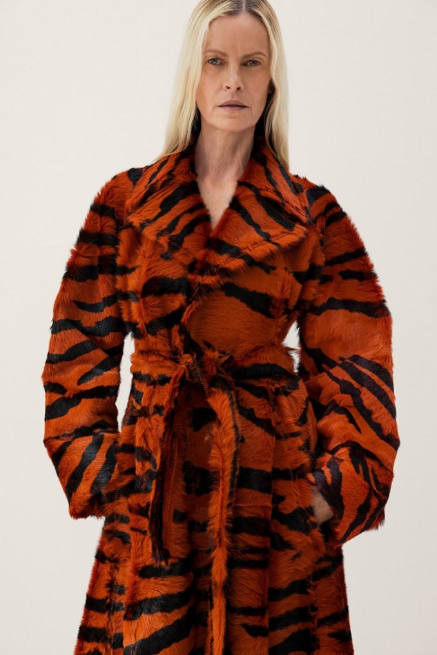 A woman with blonde hair stands indoors wearing an orange and black tiger-striped coat. The coat is tied at the waist.