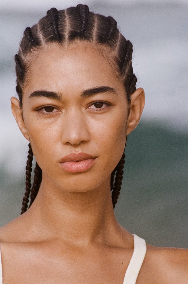 Close-up portrait of an adult woman with dark hair in cornrow braids, light skin, neutral expression, against a blurred ocean background.