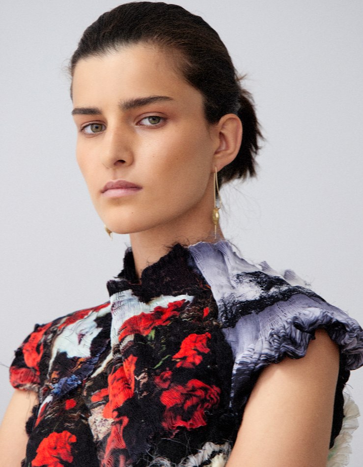 Close-up portrait of a woman with dark hair wearing an abstract floral top with earrings, posing against a light grey backdrop.