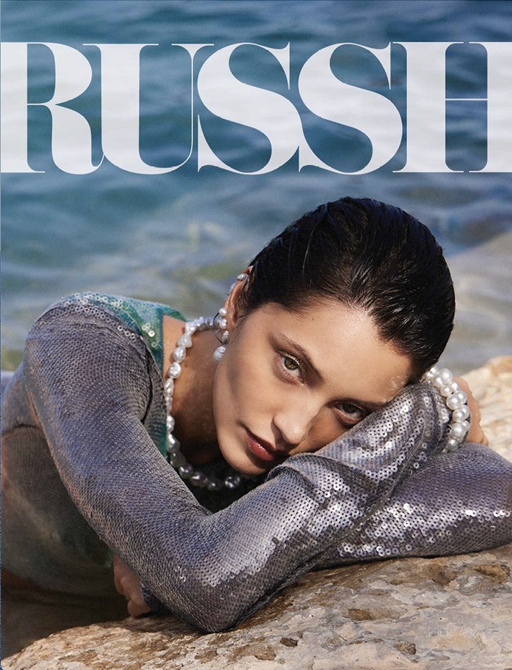 Magazine cover featuring a model in a sequined top and pearl jewelry, lying on a rock near blue water, with 'RUSSI' text overlay.