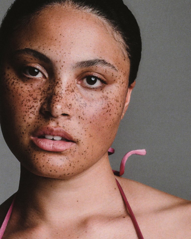 Close-up portrait of a young adult woman with freckles wearing a mauve top, posing in front of a solid grey background, beauty shot.