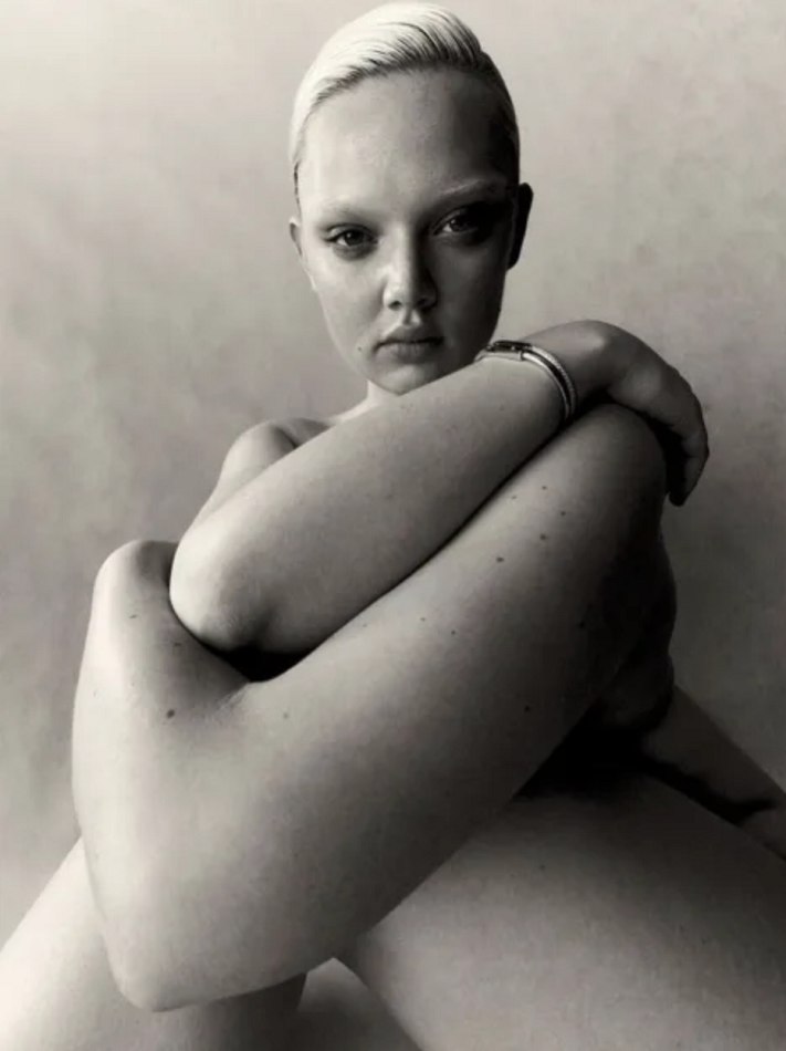 Monochrome portrait of a young adult woman with a shaved head, fair complexion, and bare shoulders posing against a light background.