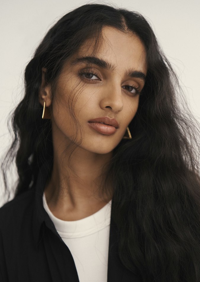 Portrait of an adult woman with long dark wavy hair, wearing gold geometric earrings, a white t-shirt and a black jacket.