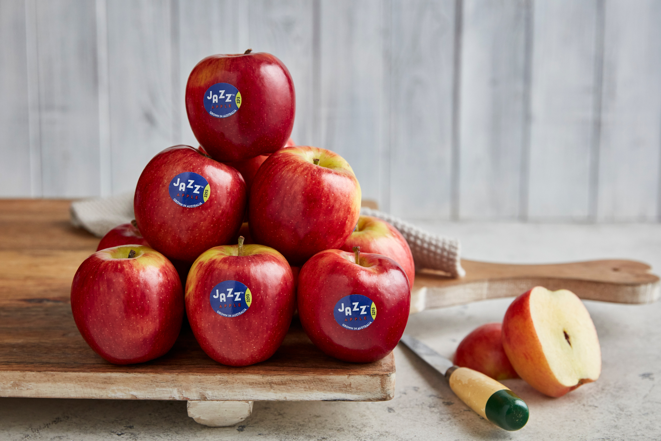 Pyramid stack of red Jazz apples with labels on a wooden cutting board next to a knife, and half an apple on a grey counter.
