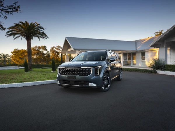 A modern gray Kia Carnival minivan is parked on a black asphalt driveway in front of a light gray house under a clear blue sky.