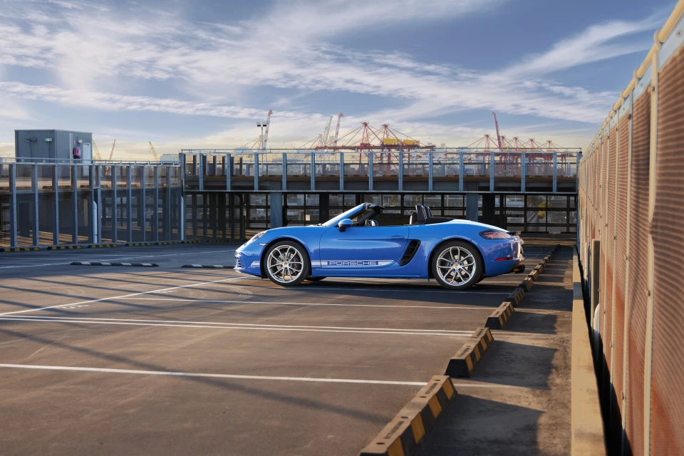A blue Porsche Boxster parked on a rooftop parking deck with a cityscape and cranes visible in the background on a sunny day.