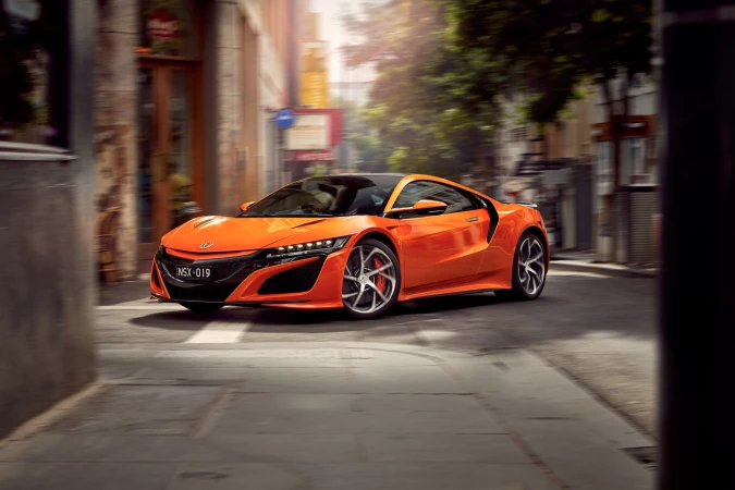 An orange 2020 Acura NSX sports car races down an urban street, showcasing its modern design and blurred motion against a backdrop of buildings.