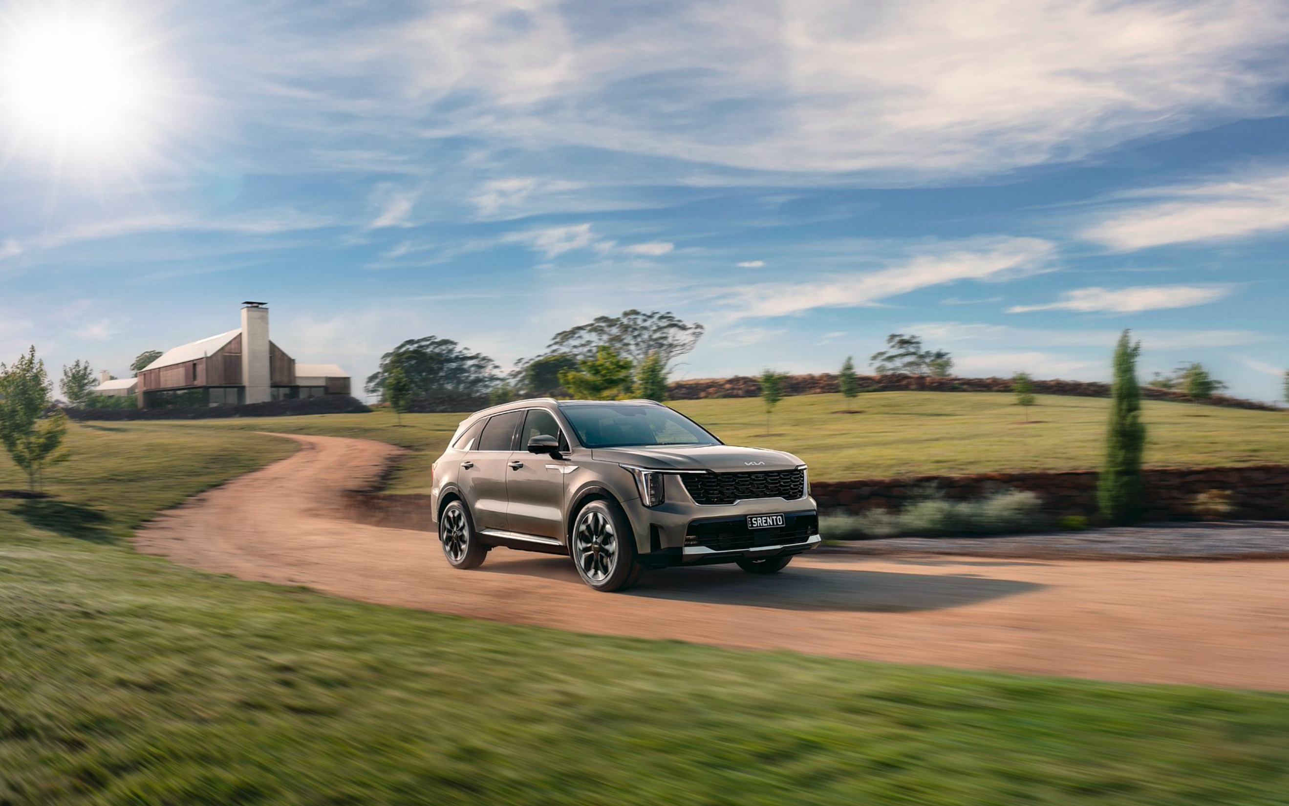Kia Sorento SUV driving on dirt road through a green grassy field with modern house and blue sky, sunny day.