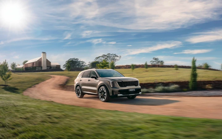 Kia Sorento SUV driving on dirt road through a green grassy field with modern house and blue sky, sunny day.