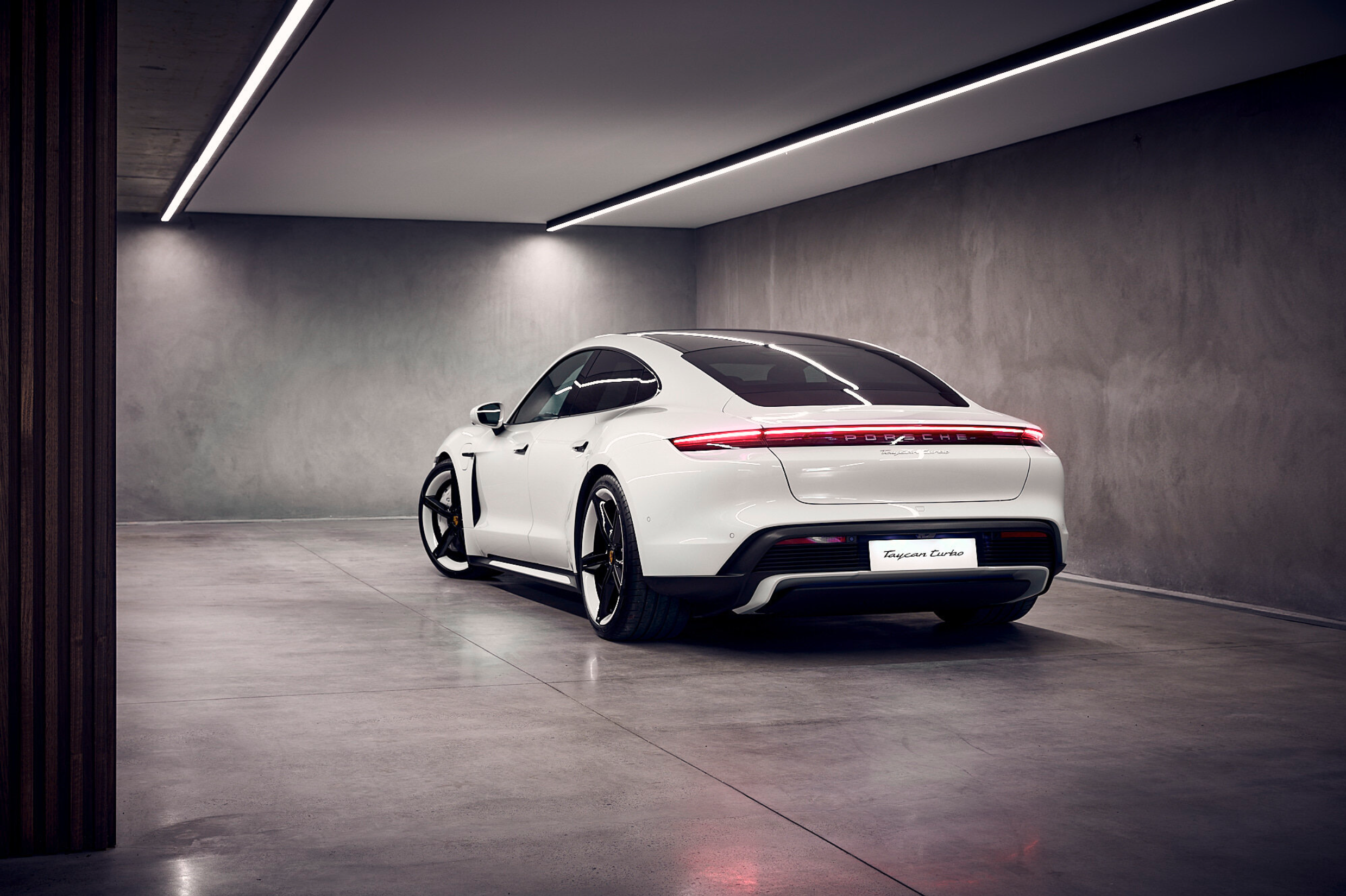 Rear view of a white Porsche Taycan Turbo parked in a garage with bright overhead lighting and a smooth concrete floor.